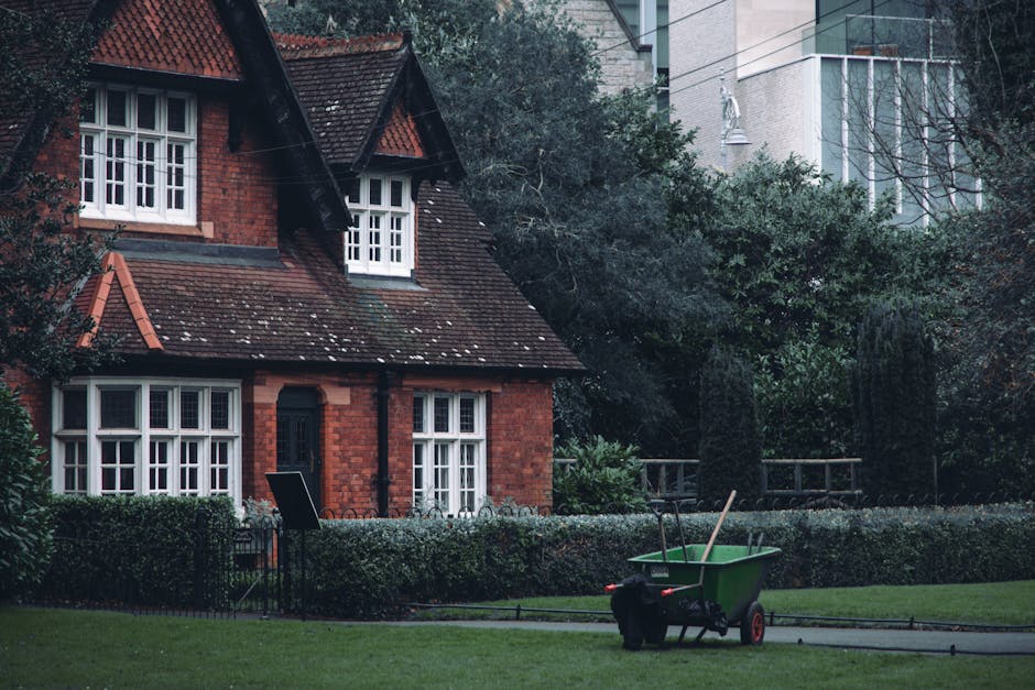 A red brick house with white-framed bay windows and a pitched roof with dark tiles, situated in a well-maintained garden. The garden features a neatly trimmed hedge along the front, lush green grass,
